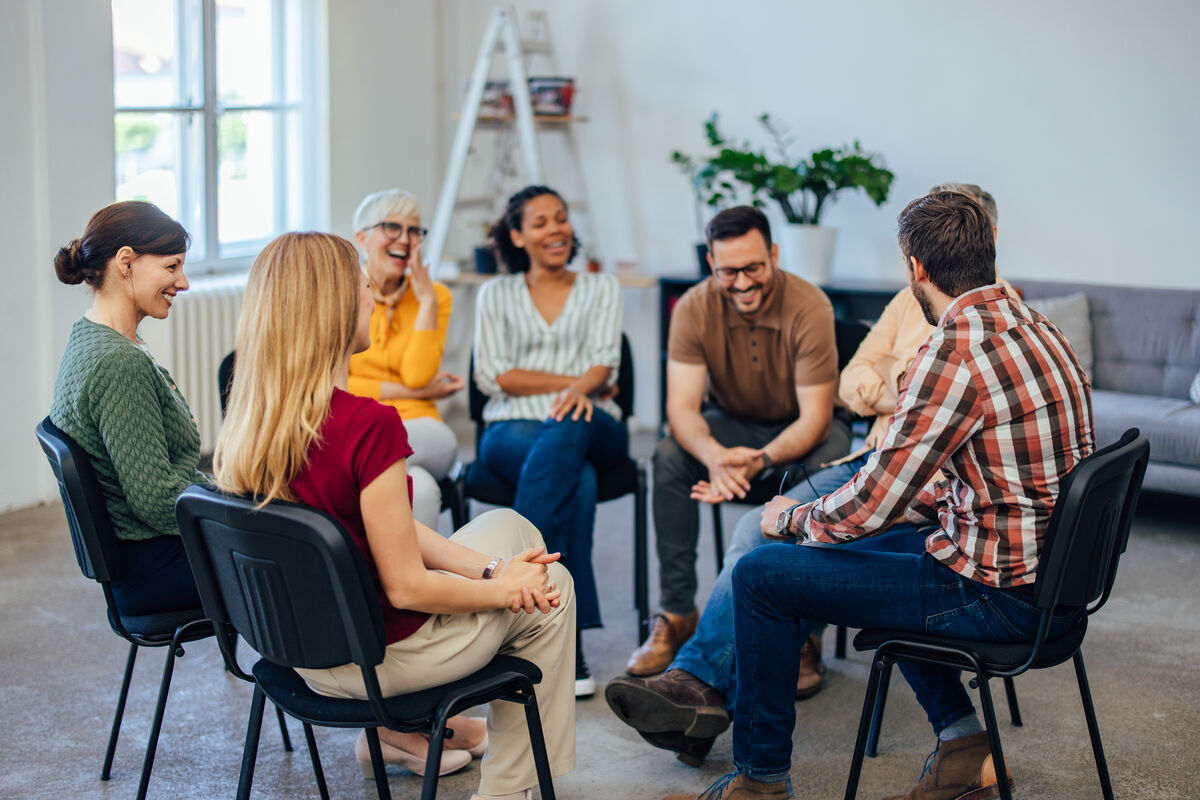 Adults in a bright room sitting in a circle, listening to a member who is speaking
